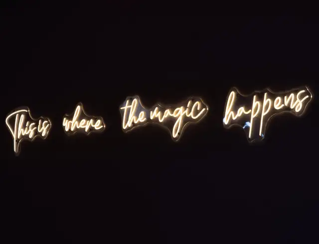 Neon sign on a dark wall reading 'This is where the magic happens'
